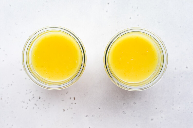 Overhead view of 2 glass jars of bright yellow vegan lemon curd on a white marble background.