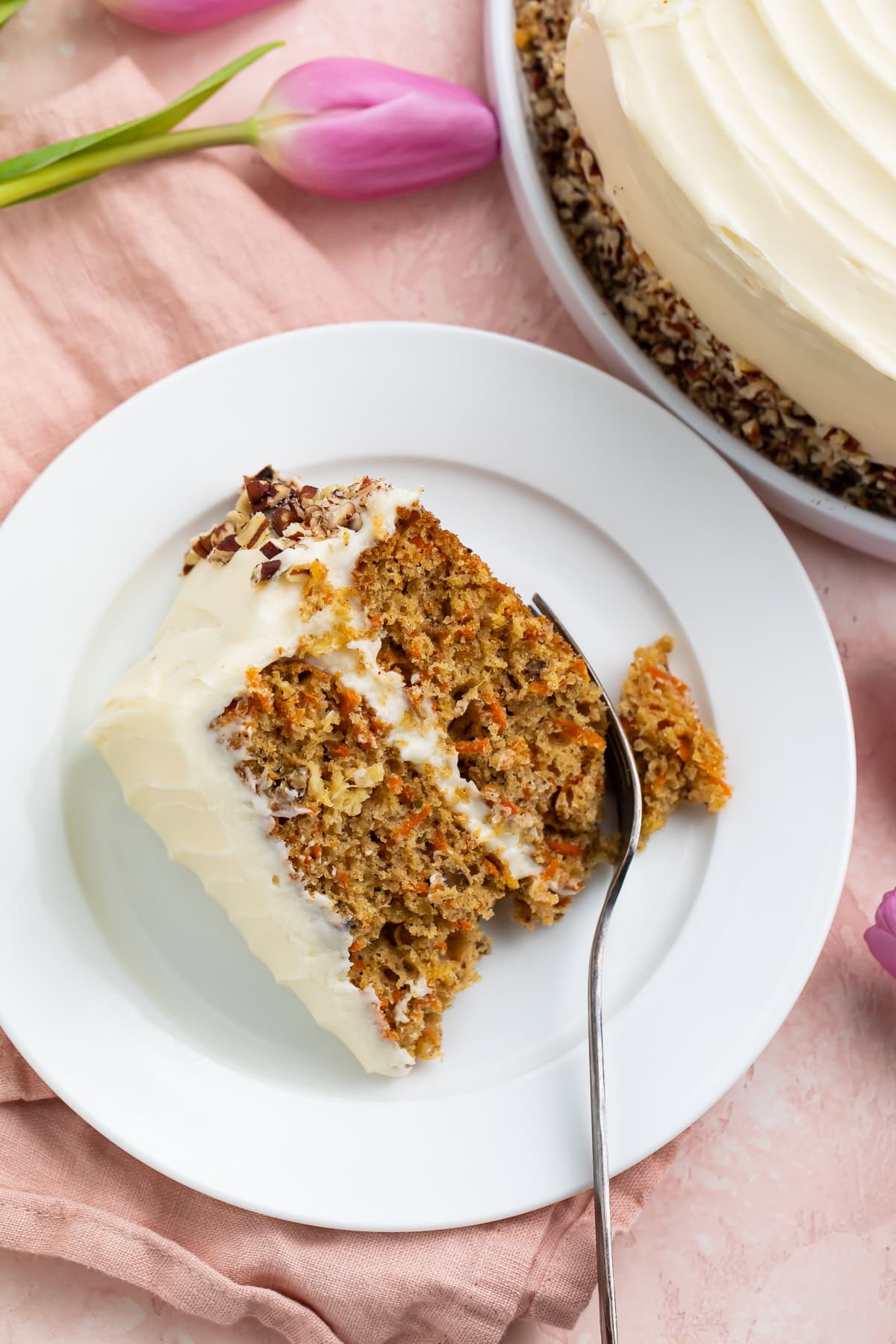 Overhead photo of a slice of frosted carrot cake on a white plate with a fork taking a bite out of one corner.
