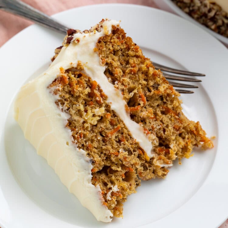 Close up of a piece of frosted carrot cake on a white plate with the edge of the cake and a pink tulip in the background.