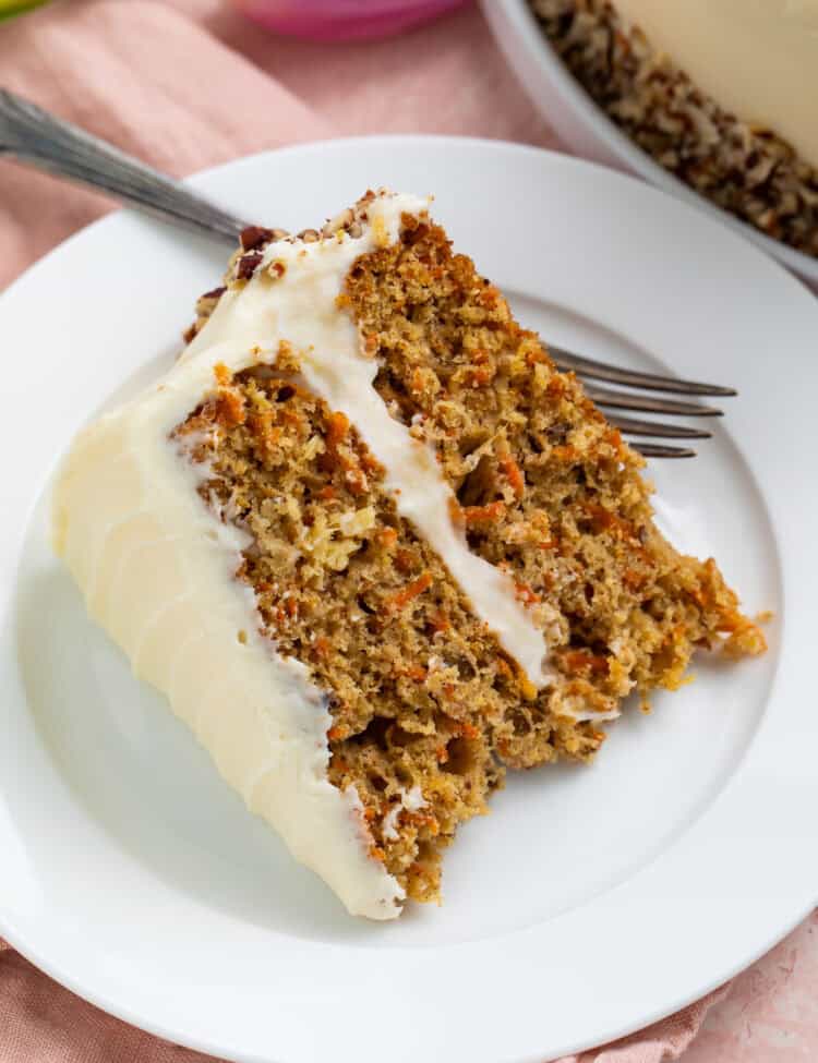 Close up of a piece of frosted carrot cake on a white plate with the edge of the cake and a pink tulip in the background.