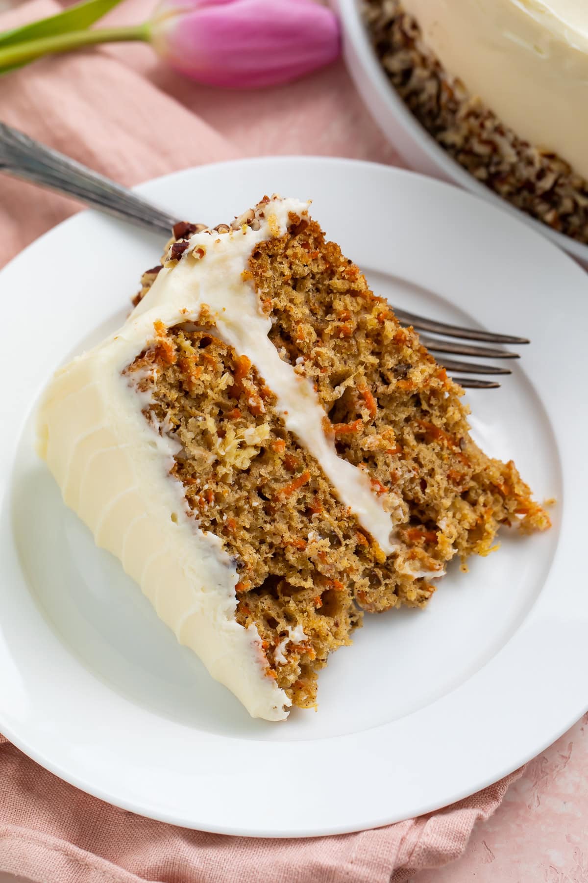 Close up of a piece of frosted carrot cake on a white plate with the edge of the cake and a pink tulip in the background.