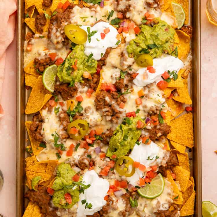 Sheet pan of nachos topped with refried beans, sour cream, cheese, guacamole and salsa.