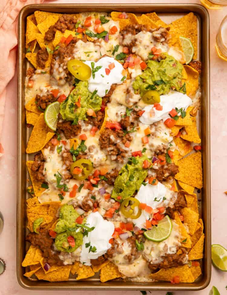 Sheet pan of nachos topped with refried beans, sour cream, cheese, guacamole and salsa.