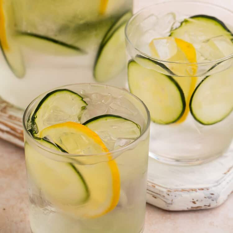 Close up of two glasses of cucumber water with lemon wheels. The jug of infused water is in the background.
