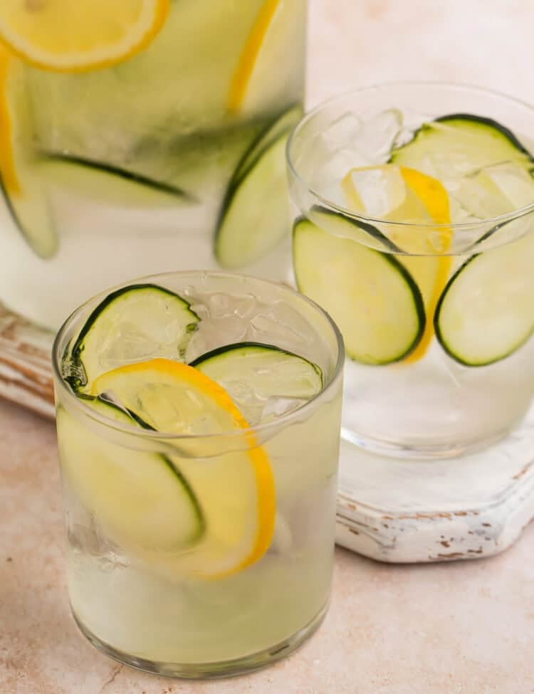 Close up of two glasses of cucumber water with lemon wheels. The jug of infused water is in the background.