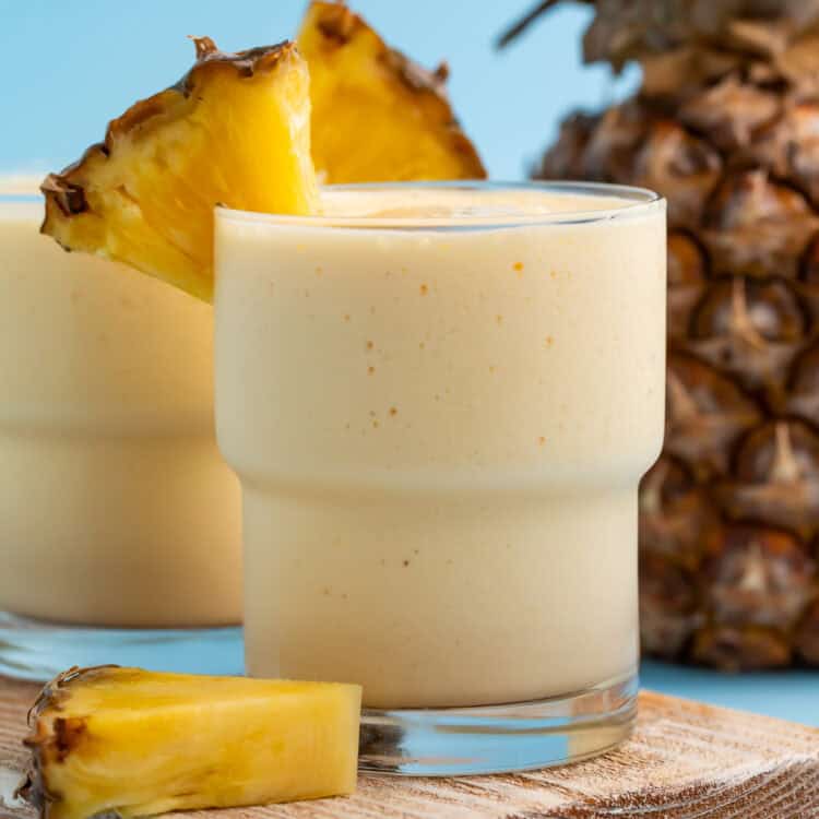 Side on view of a glass of mango pineapple smoothie on a wooden board with fresh pineapple in the background.