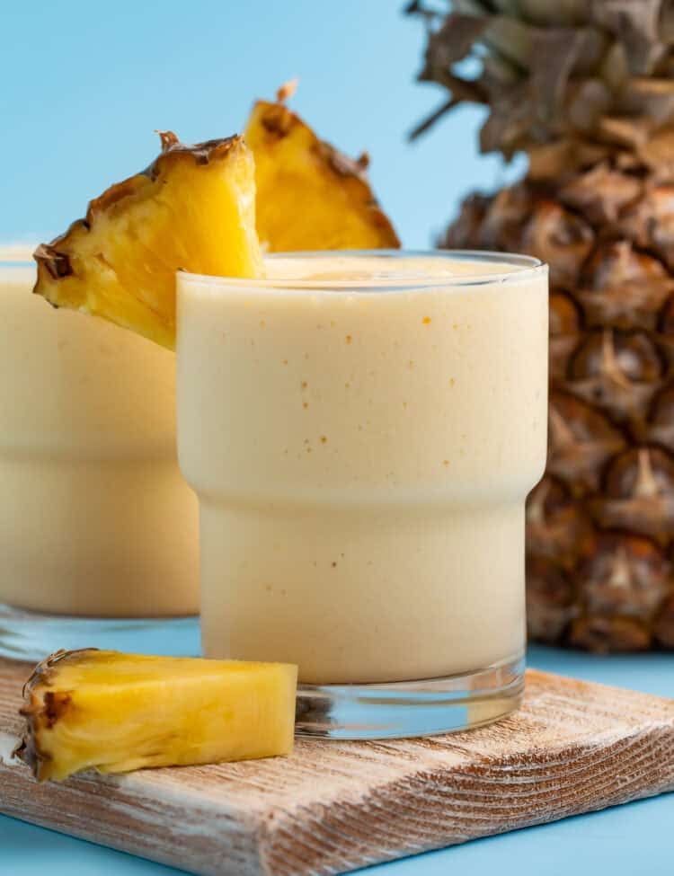 Side on view of a glass of mango pineapple smoothie on a wooden board with fresh pineapple in the background.