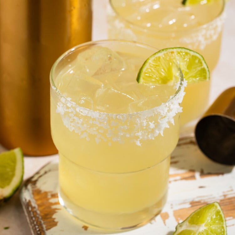 Closeup of a pair of smoke margaritas with lime wedges and a salt rim on a distressed wooden board.
