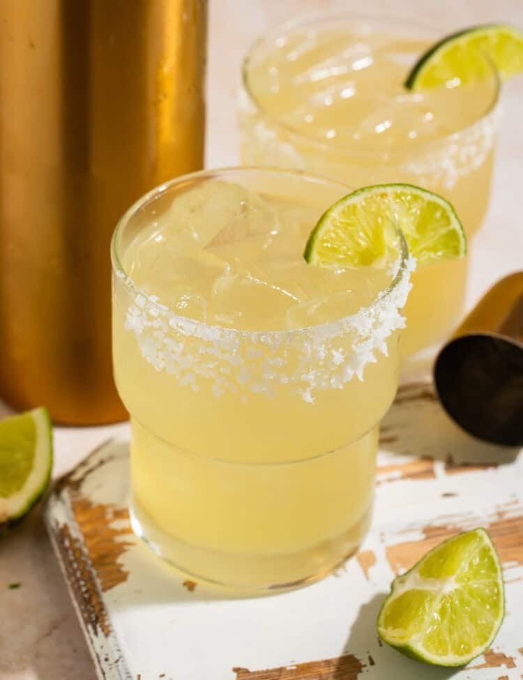 Closeup of a pair of smoke margaritas with lime wedges and a salt rim on a distressed wooden board.