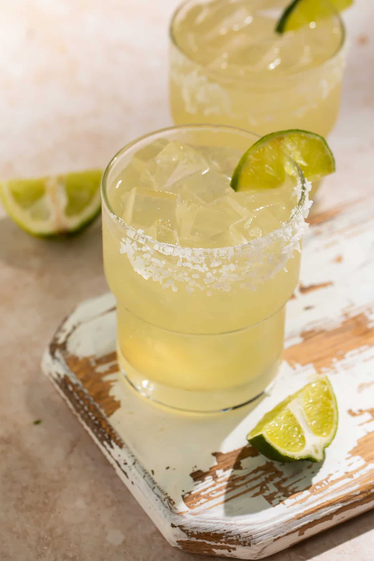 Close up of two margaritas with ice, a salt rim and lime wedges on a distressed wooden board.