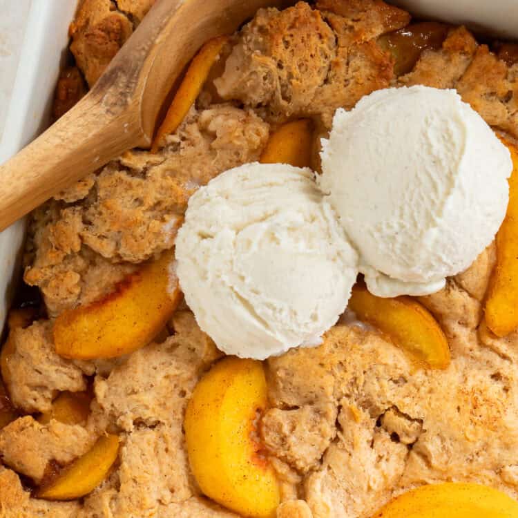 Peach cobbler in a square dish topped with vanilla ice cream. There is a wooden spoon in one side.