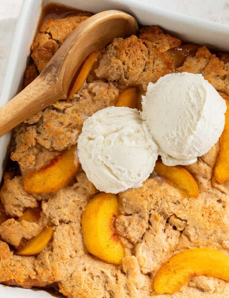 Peach cobbler in a square dish topped with vanilla ice cream. There is a wooden spoon in one side.