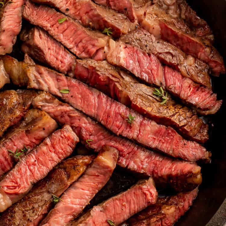 Close up of sliced medium rare rib eye steak in a cast iron skillet scattered with fresh thyme leaves.