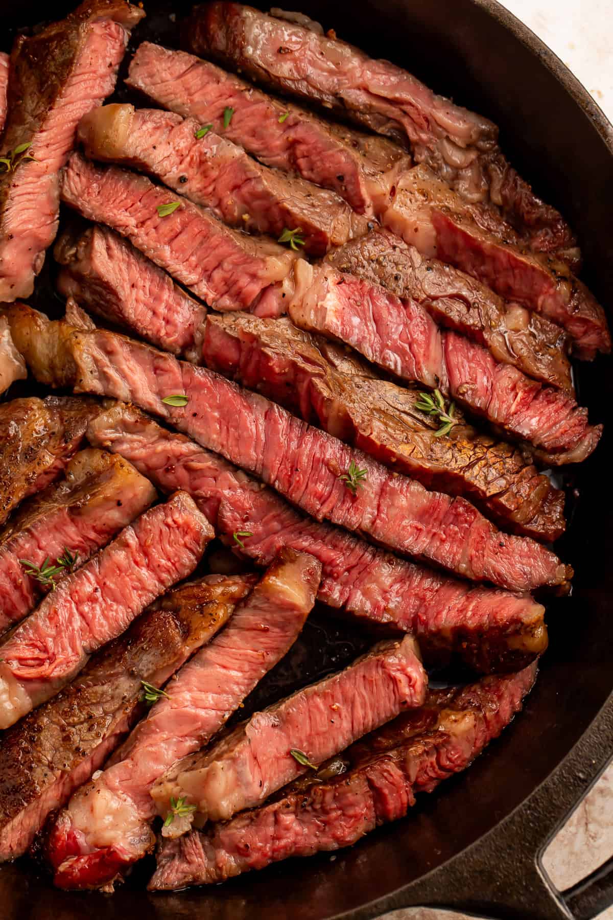 Close up of sliced medium rare rib eye steak in a cast iron skillet scattered with fresh thyme leaves.