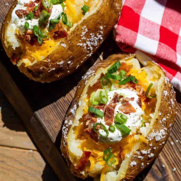 Two split open potatoes with sour cream, bacon and green onion toppings on a wooden board.