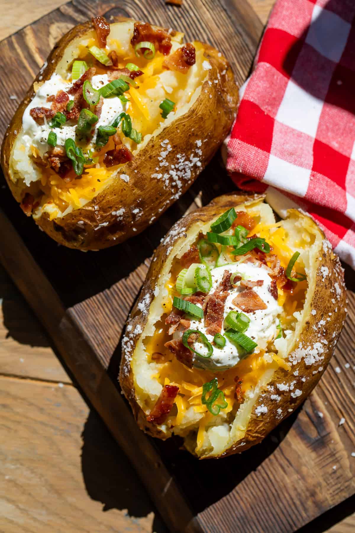 Two split open potatoes with sour cream, bacon and green onion toppings on a wooden board.