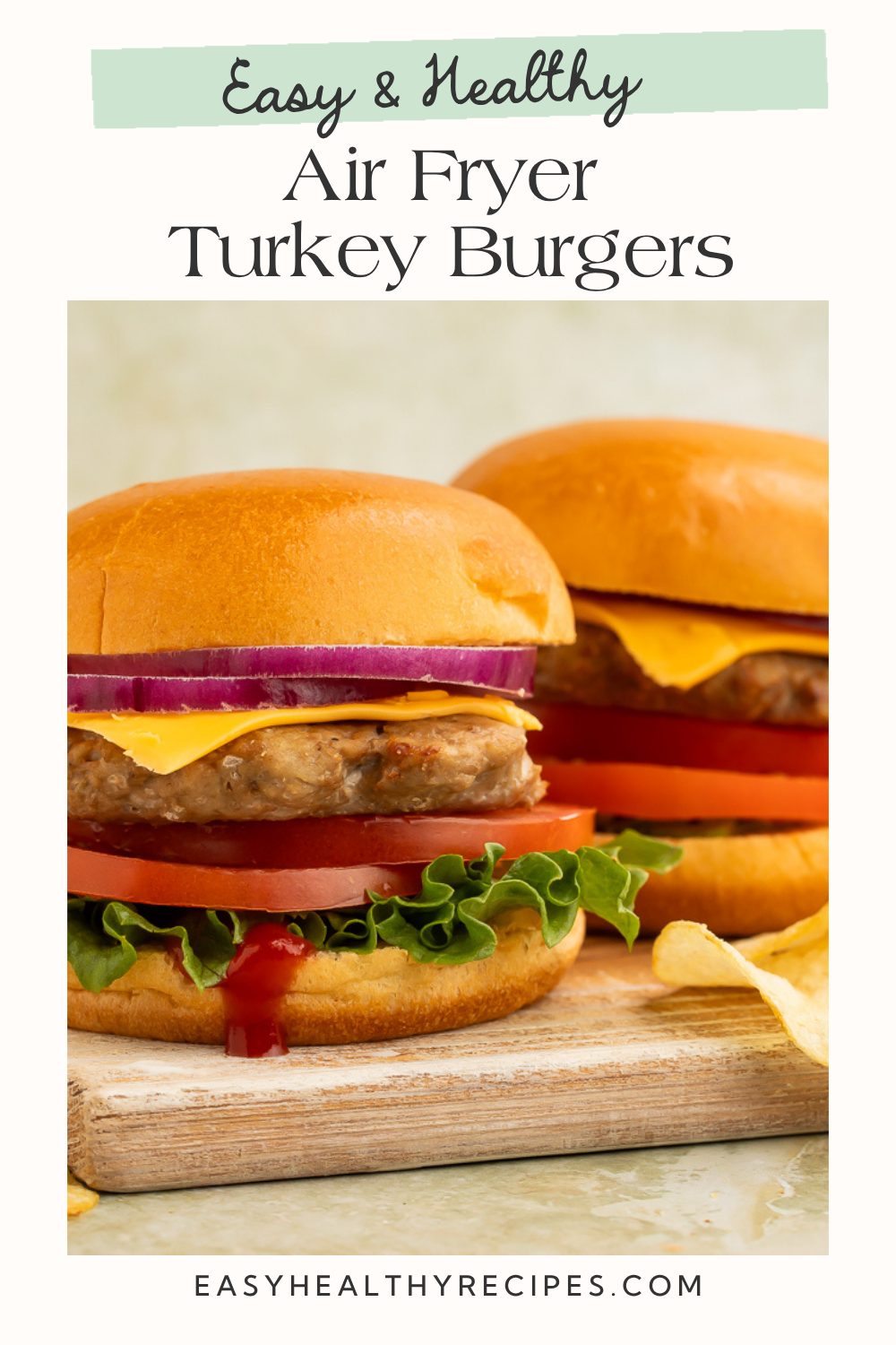 Two air fryer turkey burgers with melted cheddar, red onion, tomato, and lettuce on toasted buns, stacked on a rustic wooden cutting board with chips on the side