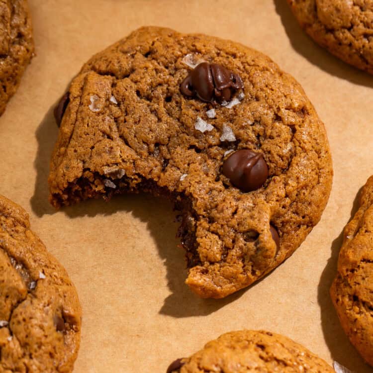 Healthy chocolate chip cookie with a bite taken out, topped with chocolate chips and flaky sea salt on parchment paper