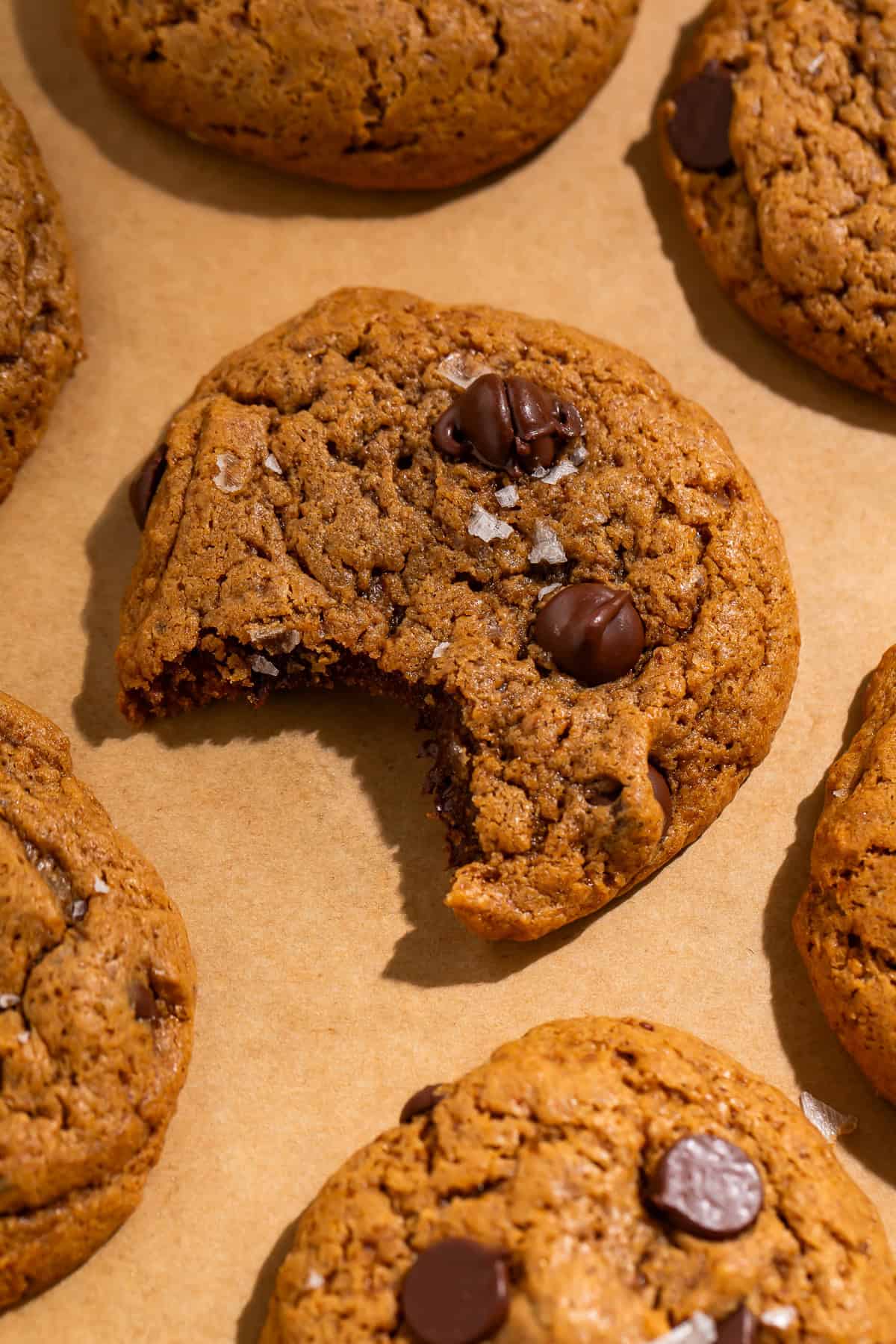 Healthy chocolate chip cookie with a bite taken out, topped with chocolate chips and flaky sea salt on parchment paper