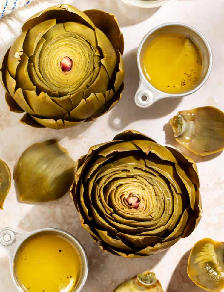 Cooked whole artichokes served with melted butter and artichoke leaves on a light countertop