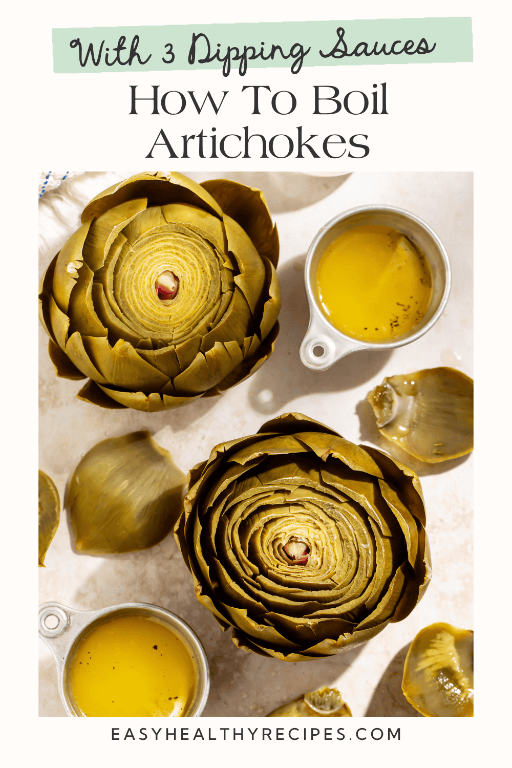 Two boiled artichokes on a white surface with small cups of golden dipping sauce