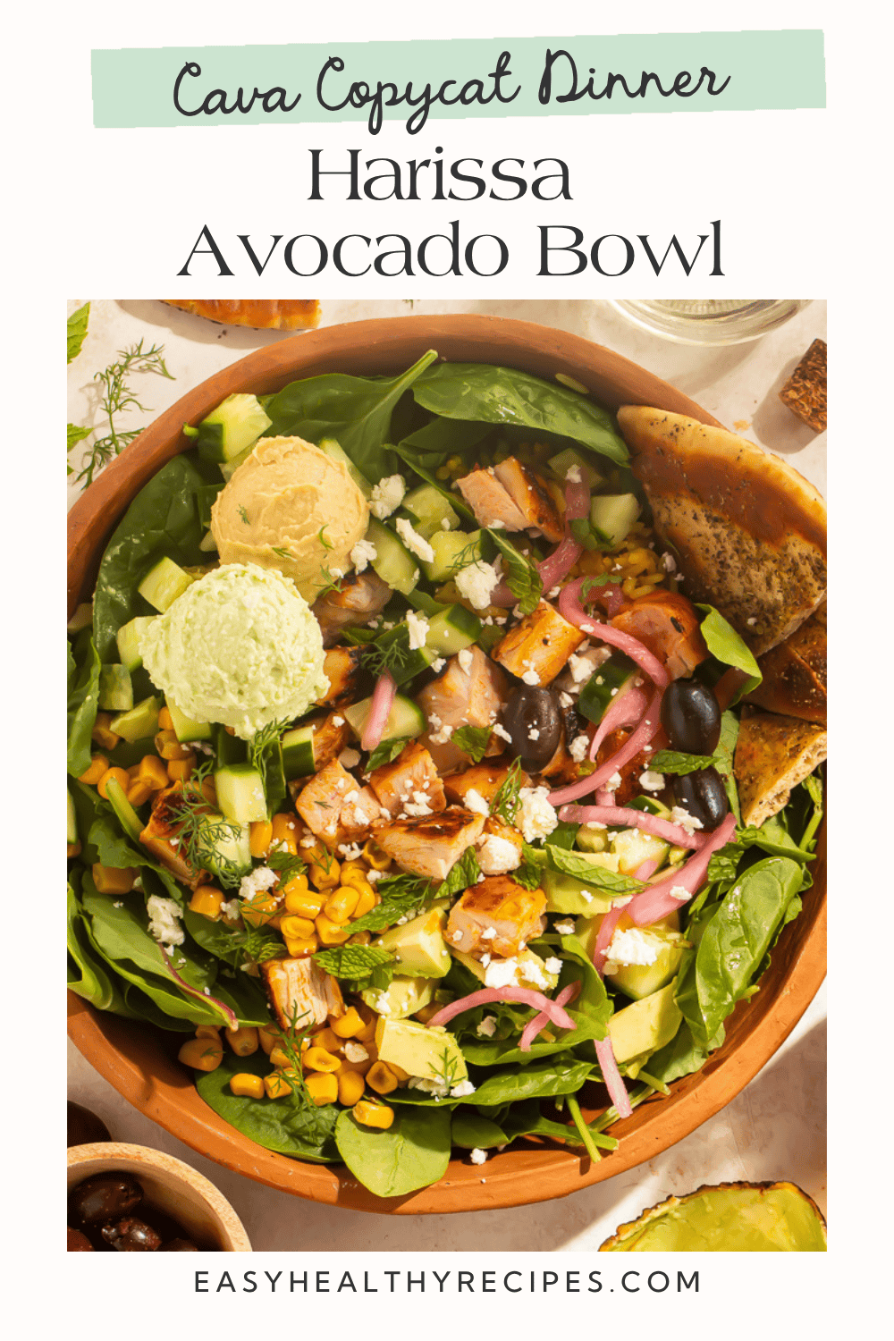 Overhead view of harissa avocado bowl with chicken, hummus, crazy feta, corn, cucumber, and pickled onions in a terracotta bowl
