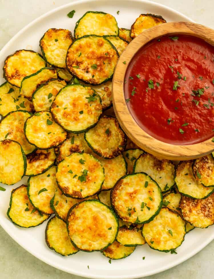 Air fryer zucchini chips served with warm marinara dipping sauce on a white platter.