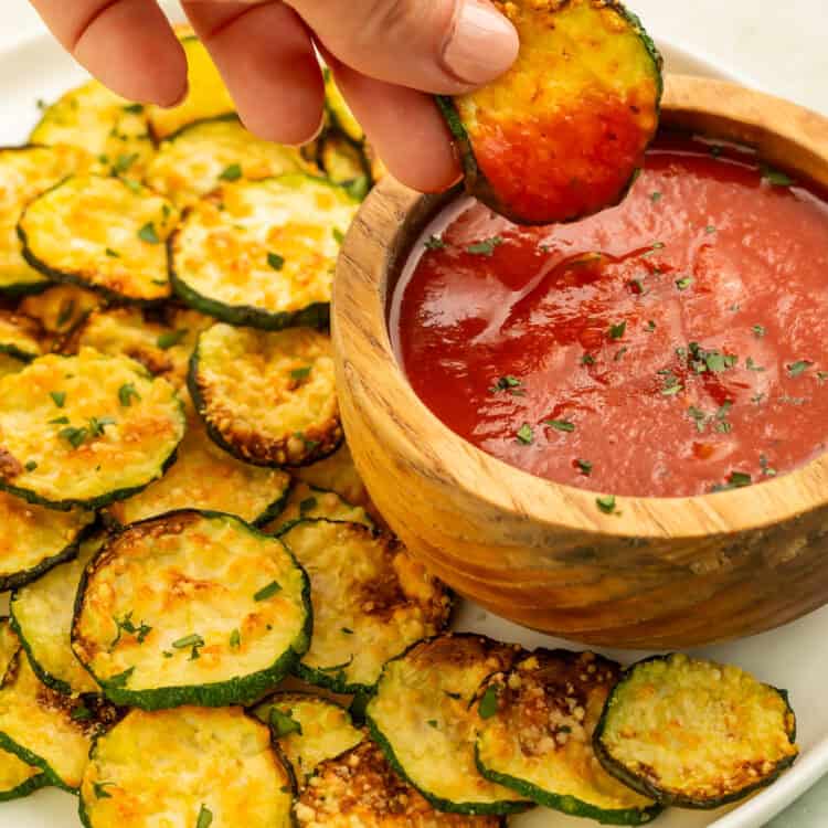 Hand dipping a crispy air fryer zucchini chip into marinara sauce.