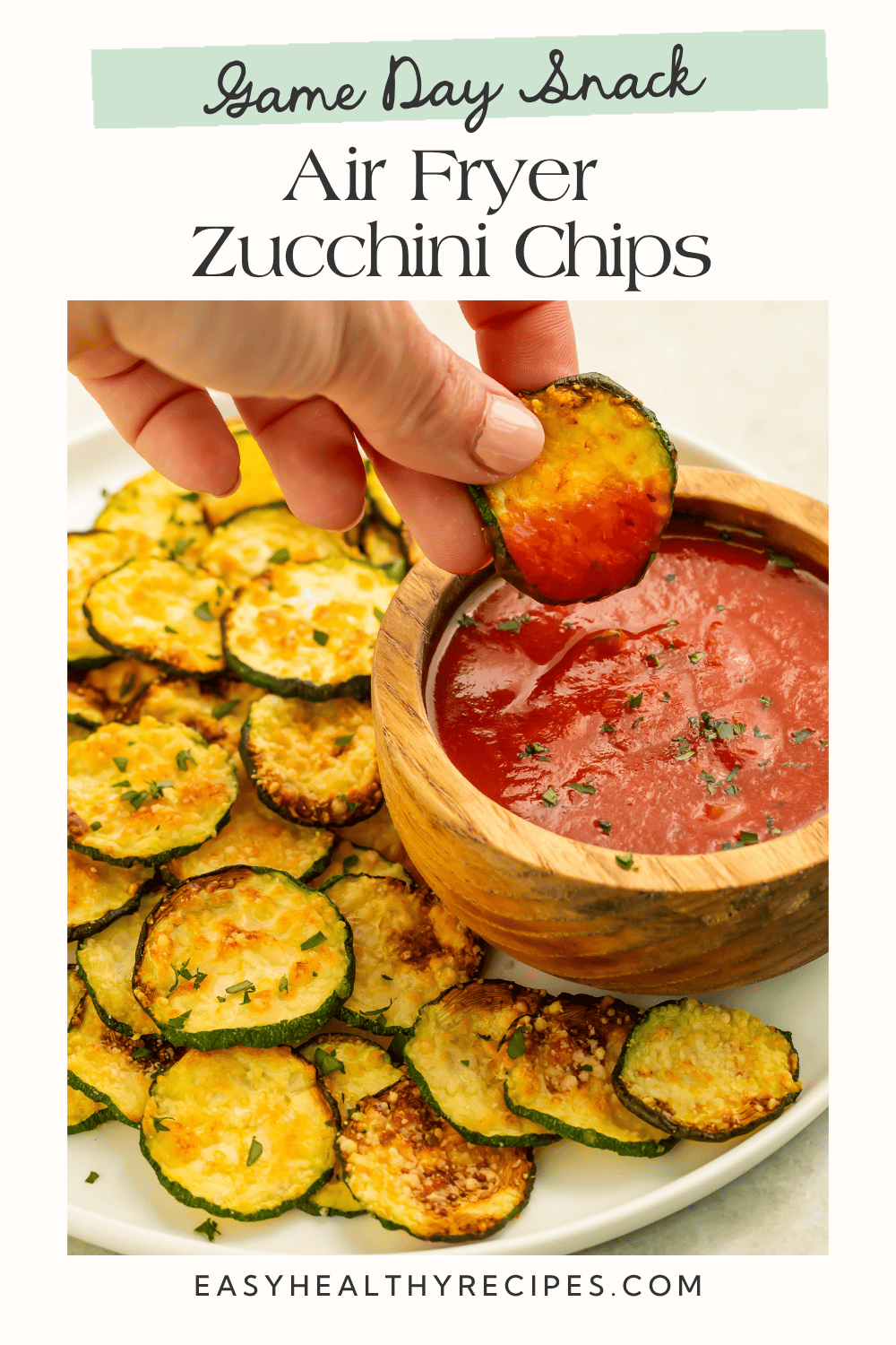 Hand dipping a crispy air fryer zucchini chip into marinara sauce beside a plate of golden parmesan zucchini chips.