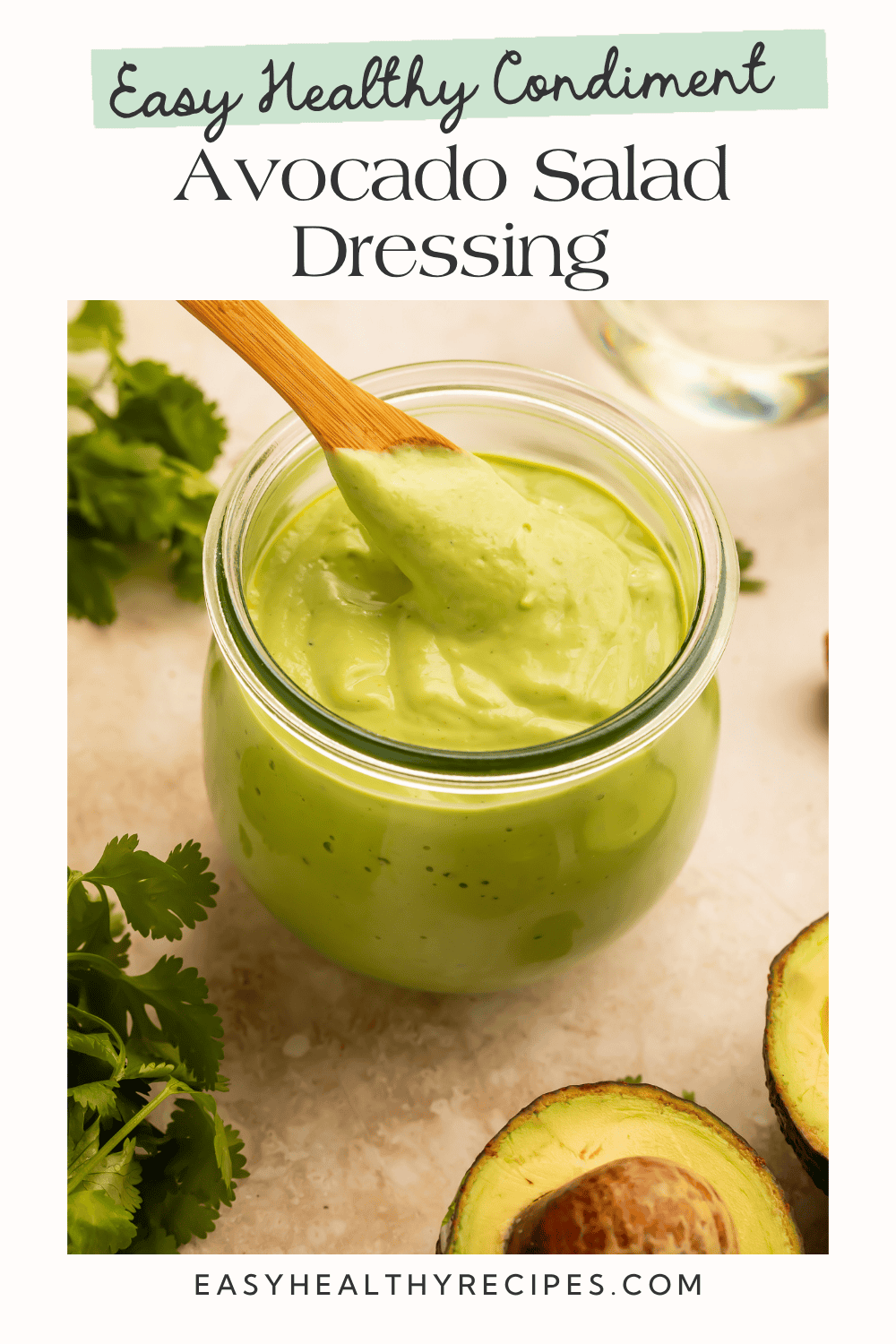 Creamy avocado salad dressing in a glass jar with a wooden spoon, surrounded by fresh cilantro and avocado halves