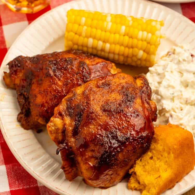 Baked BBQ chicken thighs served with potato salad, corn on the cob, and cornbread on a dinner plate.