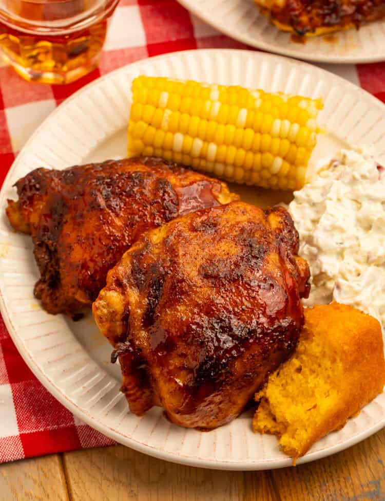 Baked BBQ chicken thighs served with potato salad, corn on the cob, and cornbread on a dinner plate.