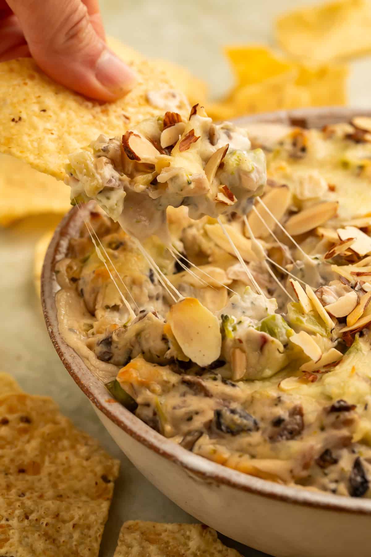 A tortilla chip scooping up creamy baked broccoli cheese dip with melted cheese, mushrooms, broccoli, and toasted sliced almonds