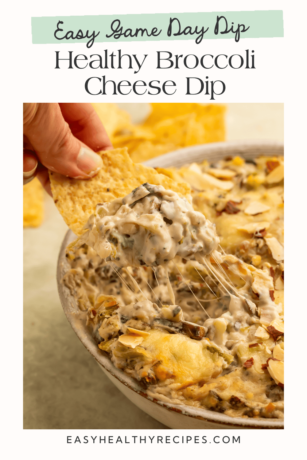 Tortilla chip scooping creamy baked broccoli cheese dip with stretchy melted cheese, broccoli, mushrooms, and toasted almonds