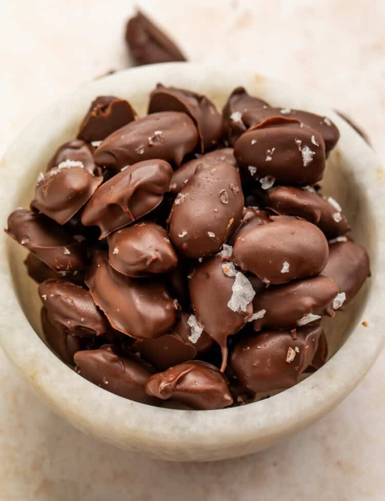 Homemade dark chocolate-covered almonds sprinkled with flaky sea salt in a white bowl.