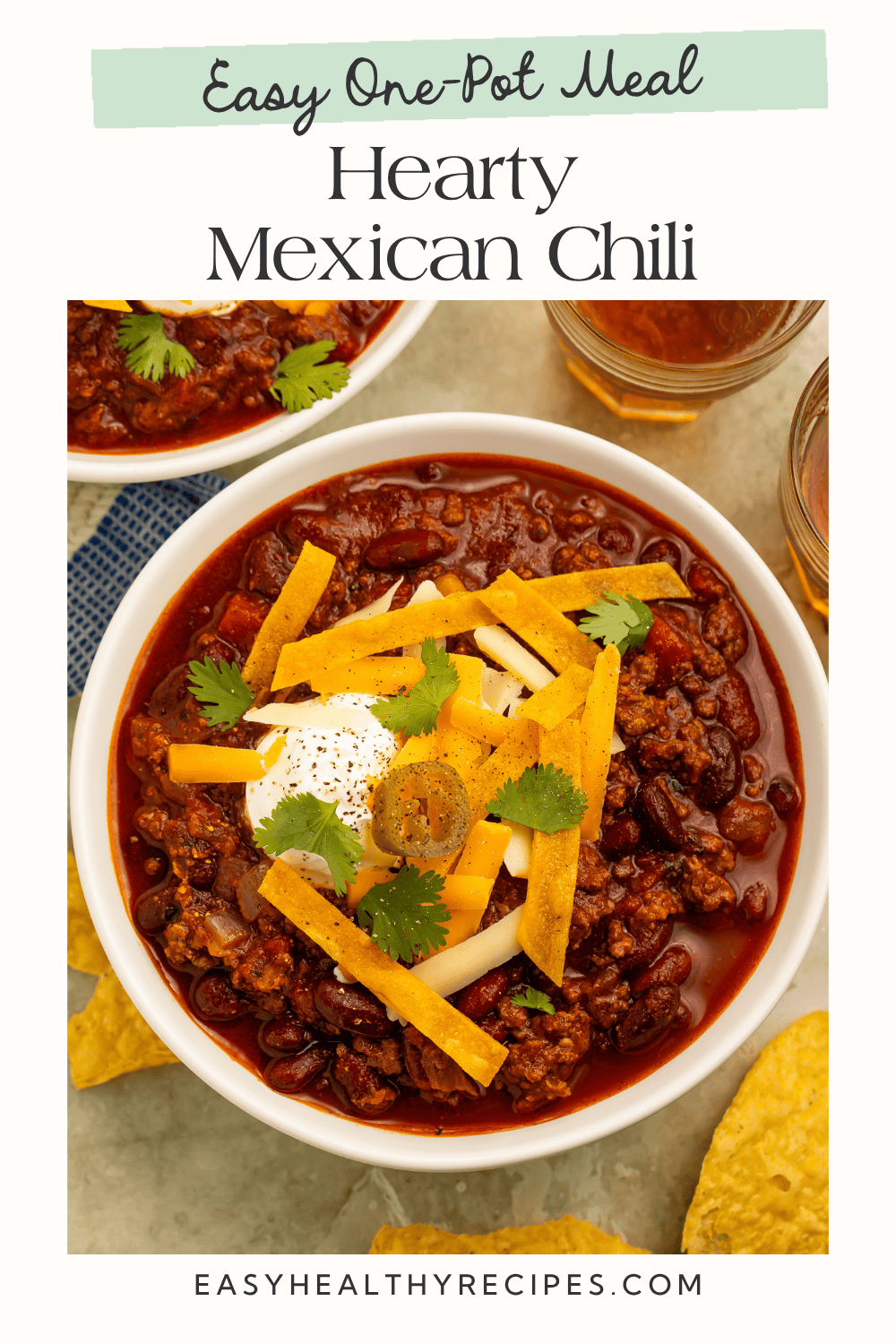 Mexican chili in a white bowl topped with tortilla strips, cheese, sour cream, cilantro, and a jalapeño slice.