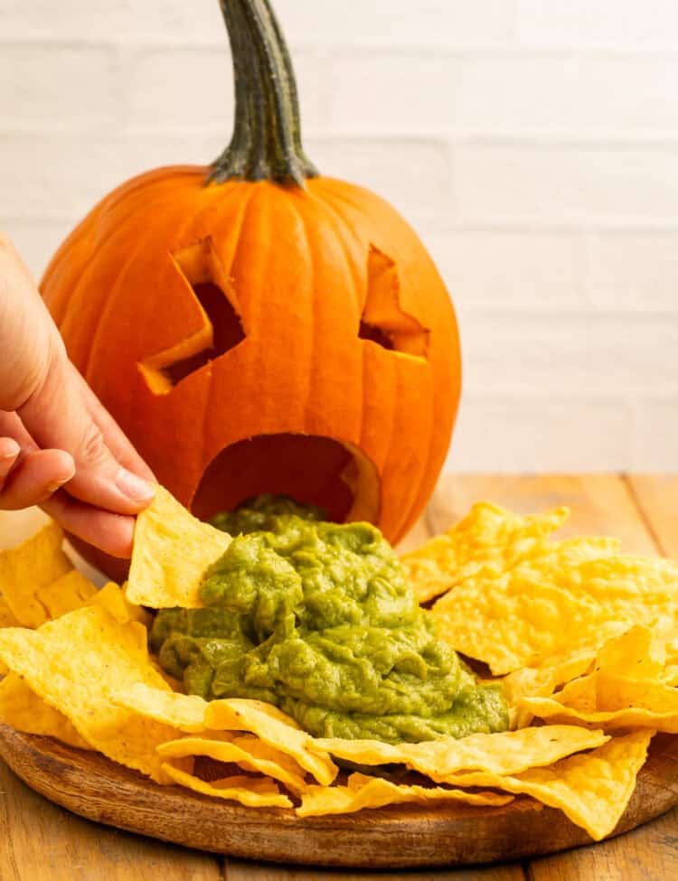 Hand dipping a tortilla chip into guacamole spilling from a carved puking pumpkin’s mouth.