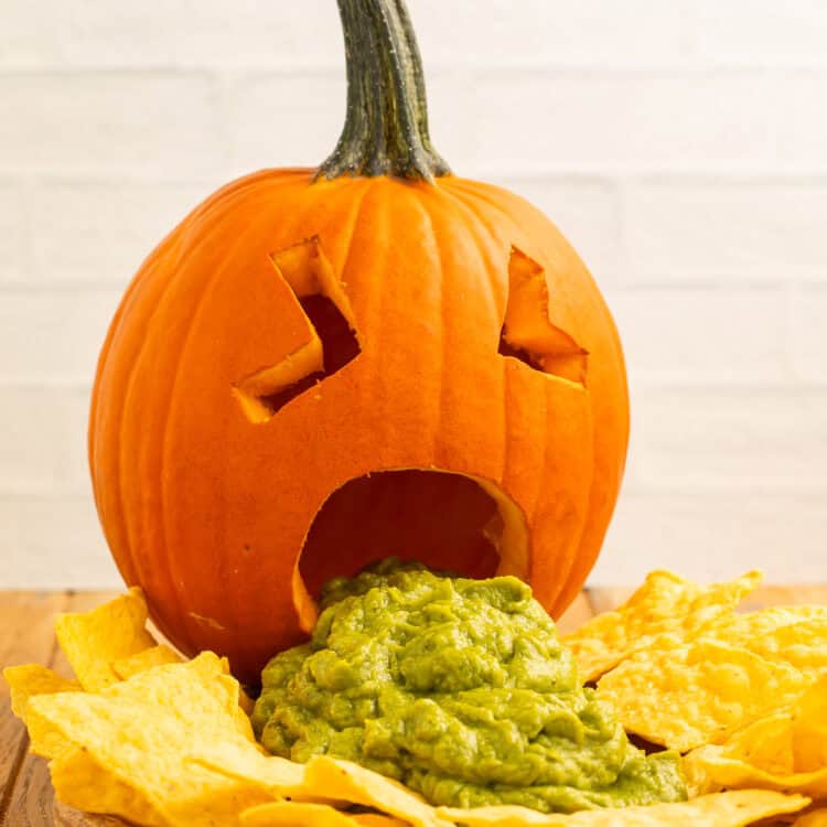 Puking pumpkin with guacamole spilling from its carved mouth, surrounded by tortilla chips on a wooden board.