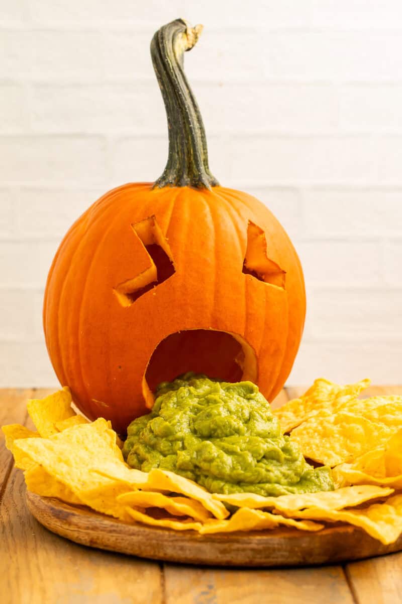 Puking pumpkin with guacamole spilling from its carved mouth, surrounded by tortilla chips on a wooden board.