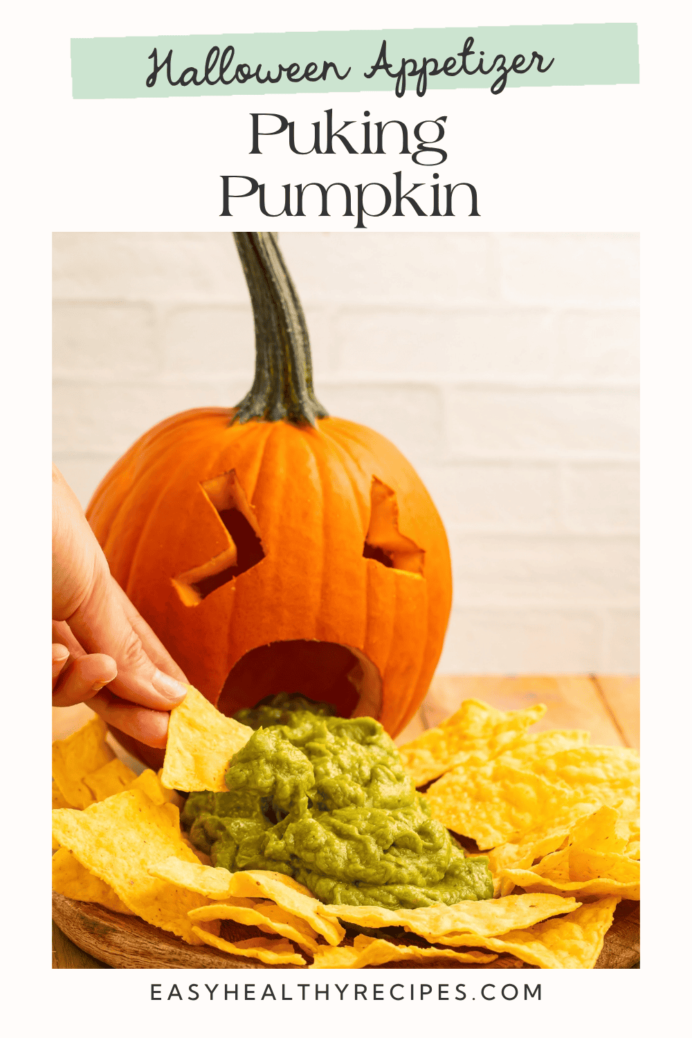 Hand dipping a tortilla chip into guacamole spilling from a carved pumpkin’s mouth.