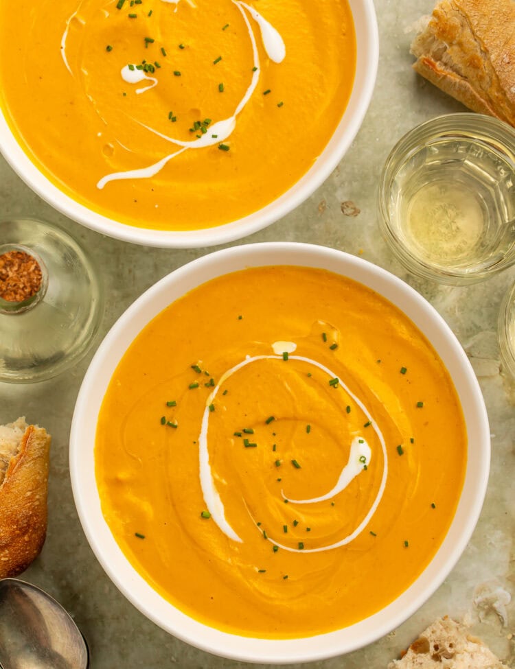 Two bowls of creamy pumpkin bisque topped with cream swirls and chopped chives, served with crusty bread.