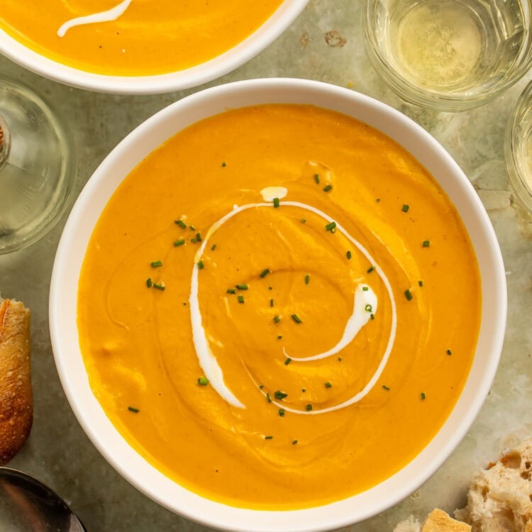 Creamy pumpkin bisque in a white bowl with a swirl of cream and chopped chives, with crusty bread and a spoon alongside.