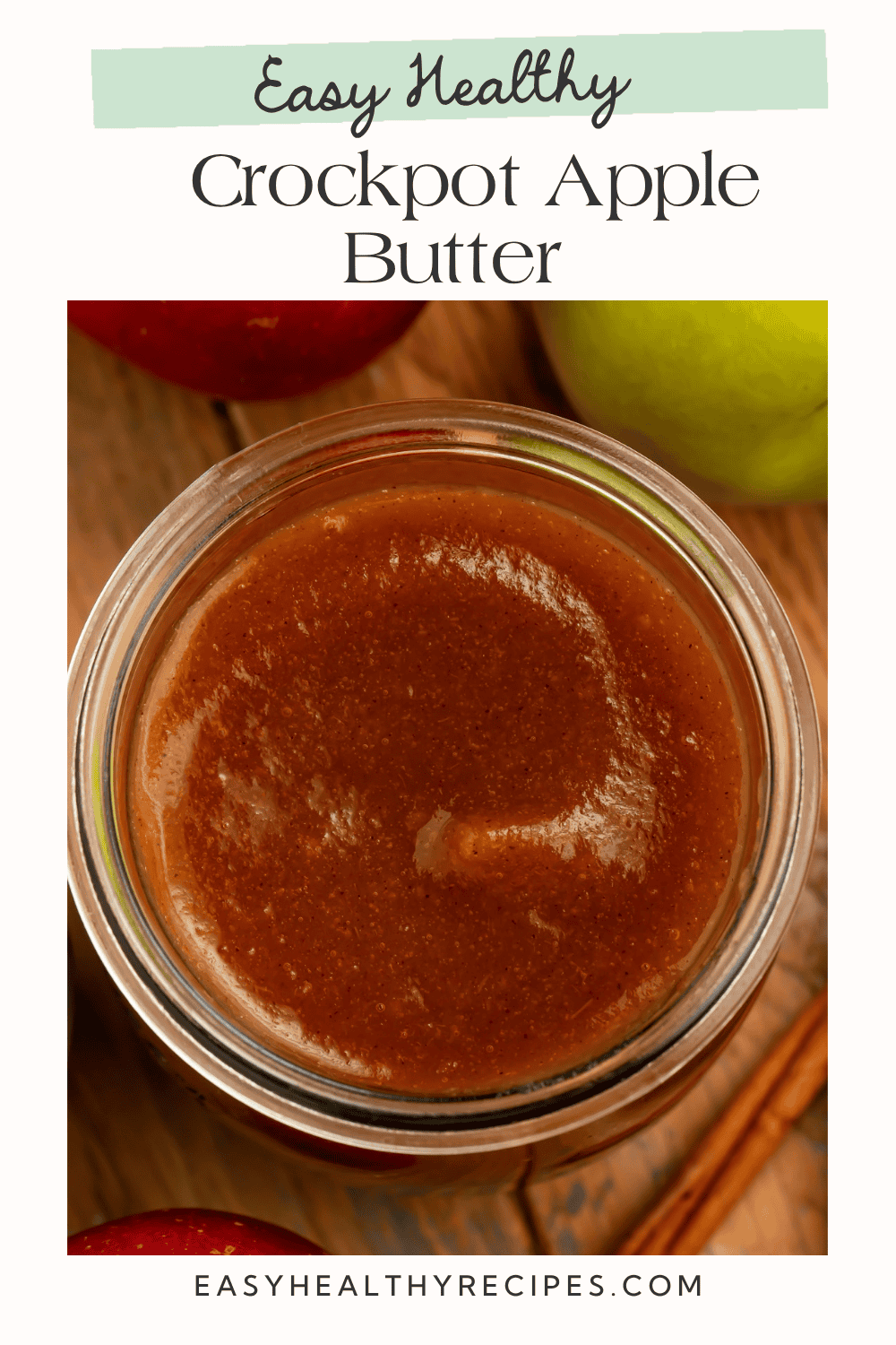 Thick homemade crockpot apple butter shown from above, with a smooth swirl and fresh red and green apples nearby.