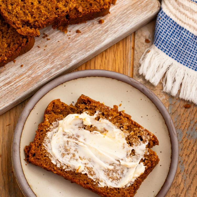 A slice of gluten-free pumpkin bread spread with butter on a ceramic plate, with more slices on a board behind it.