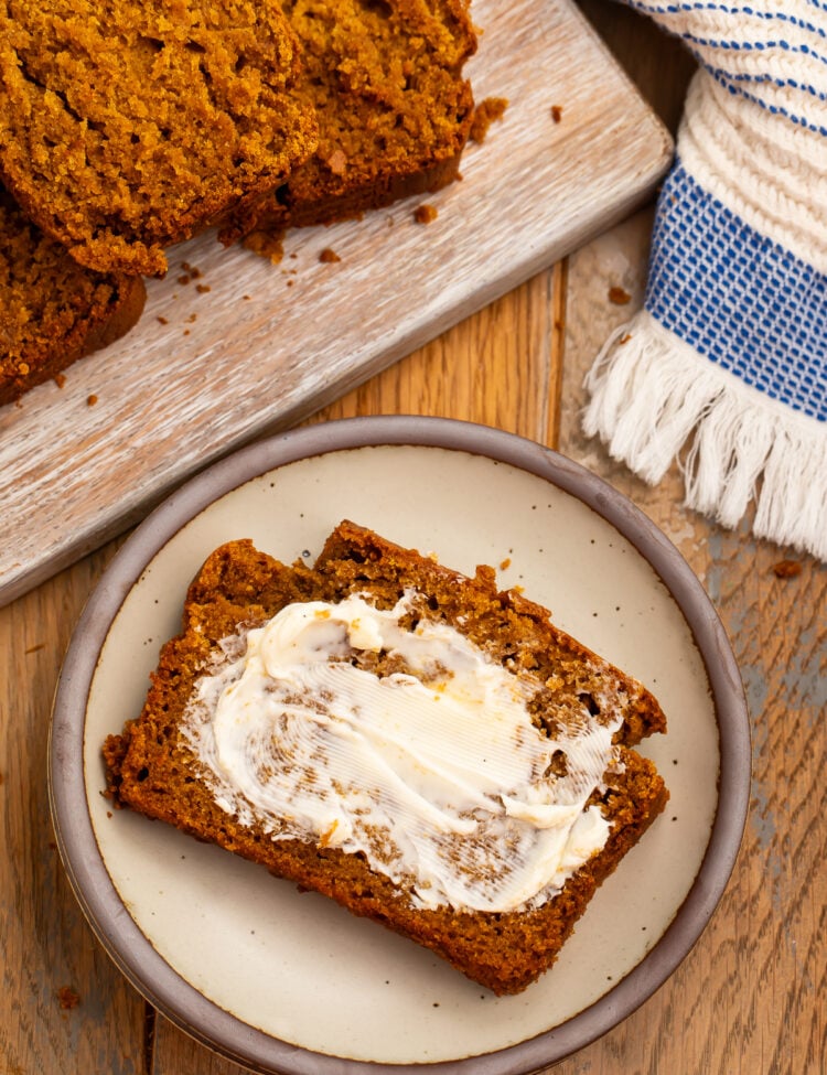 A slice of gluten-free pumpkin bread spread with butter on a ceramic plate, with more slices on a board behind it.