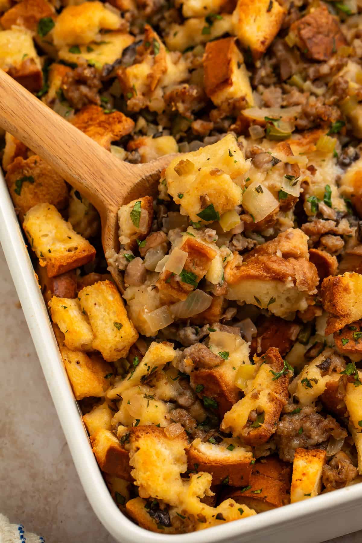 Wooden spoon lifting gluten free stuffing with browned bread cubes, sausage, mushrooms, onions, and celery from a baking dish.