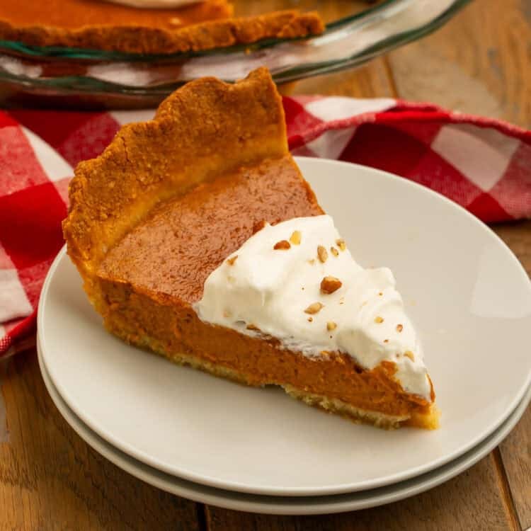 Slice of keto pumpkin pie with almond flour crust, whipped cream, and chopped nuts on a white plate.