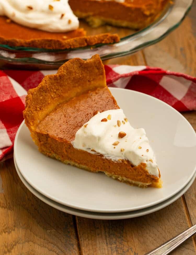 Slice of keto pumpkin pie with almond flour crust, whipped cream, and chopped nuts on a white plate.