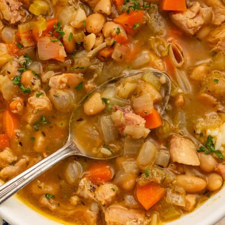 Close up of smoked turkey soup with a spoon holding turkey, white beans, and vegetables in golden broth.