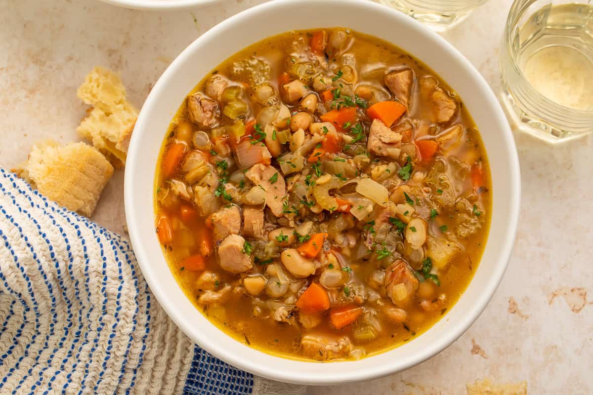 Smoked turkey soup with white beans, carrots, celery, and chopped herbs in a white bowl beside crusty bread.
